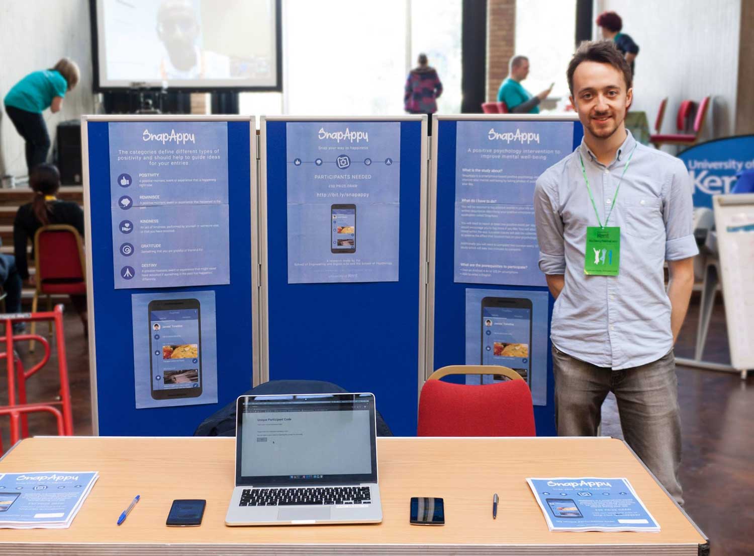 Photo of James standing at a stall with posters, leaflets and a laptop to attract participants.